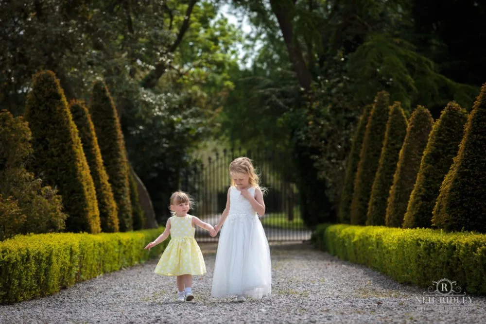 Two little girls walk hand in hand down a tree lined drive at The Great Hall of Mains.
